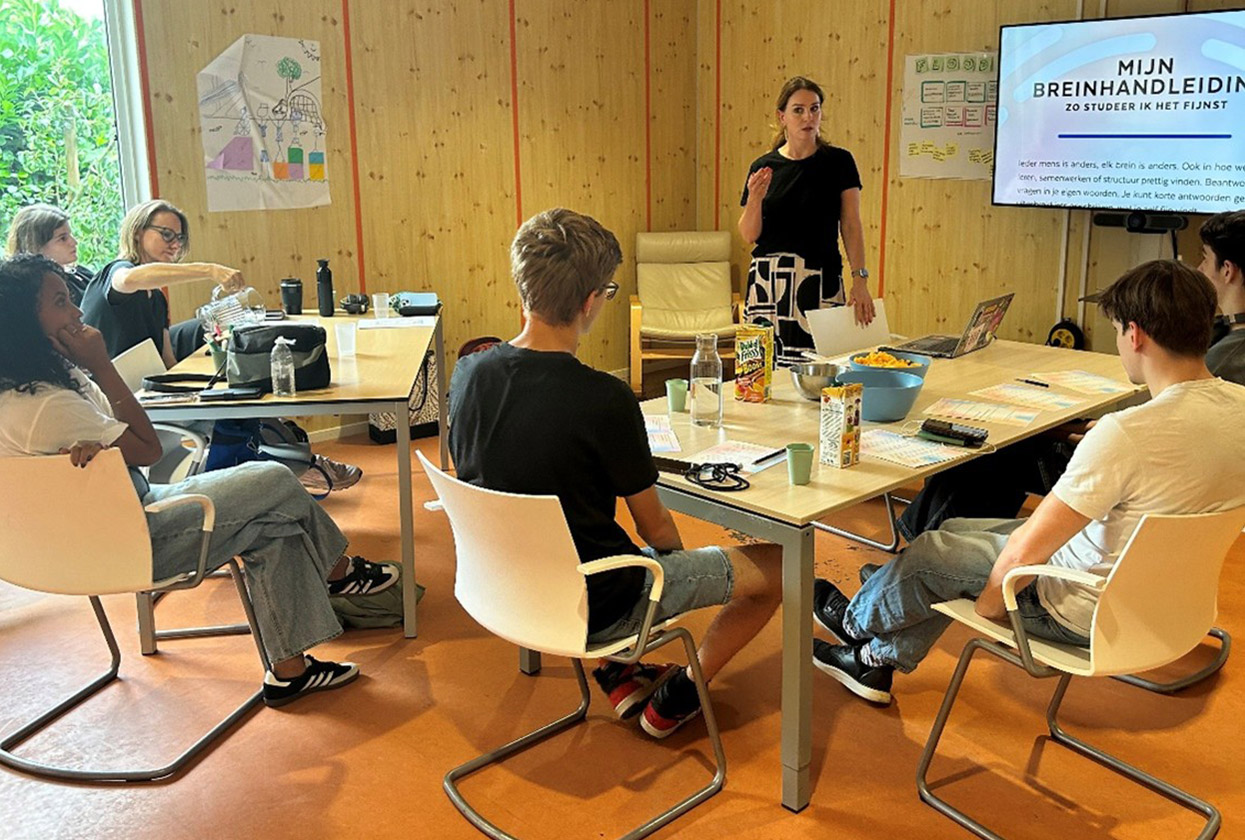 Een aantal jonge mensen aan een grote tafel, kijkend naar een docent die naast een beeldscherm een cursus geeft.